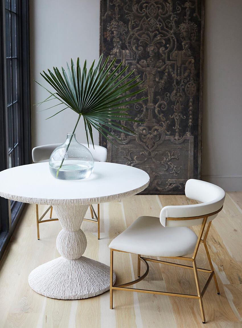 Lifestyle photo with a round white table, two white upholstery chairs with gold metal legs and supports, a birch colored plank floor, glass vase with a single stemmed fan like leaf plant, and a decorative carving artwork in the background.